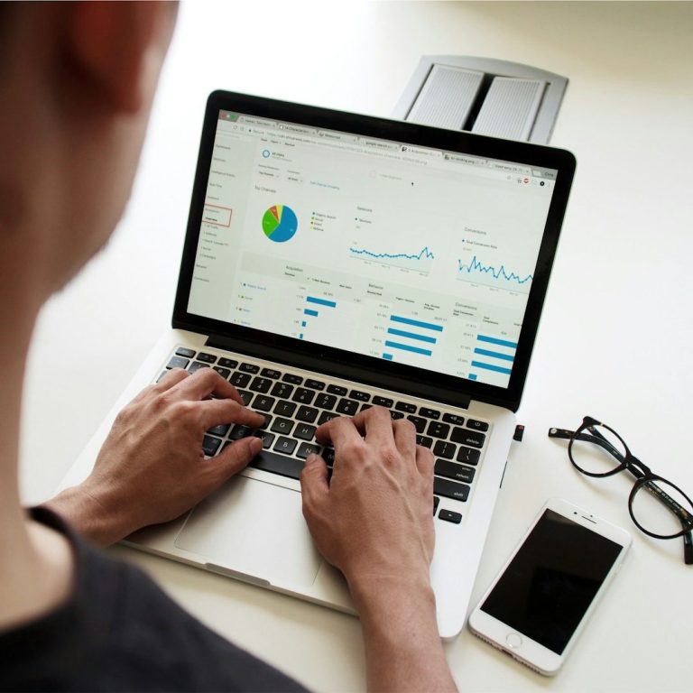 Person typing on a laptop displaying data charts, with a phone and glasses nearby.