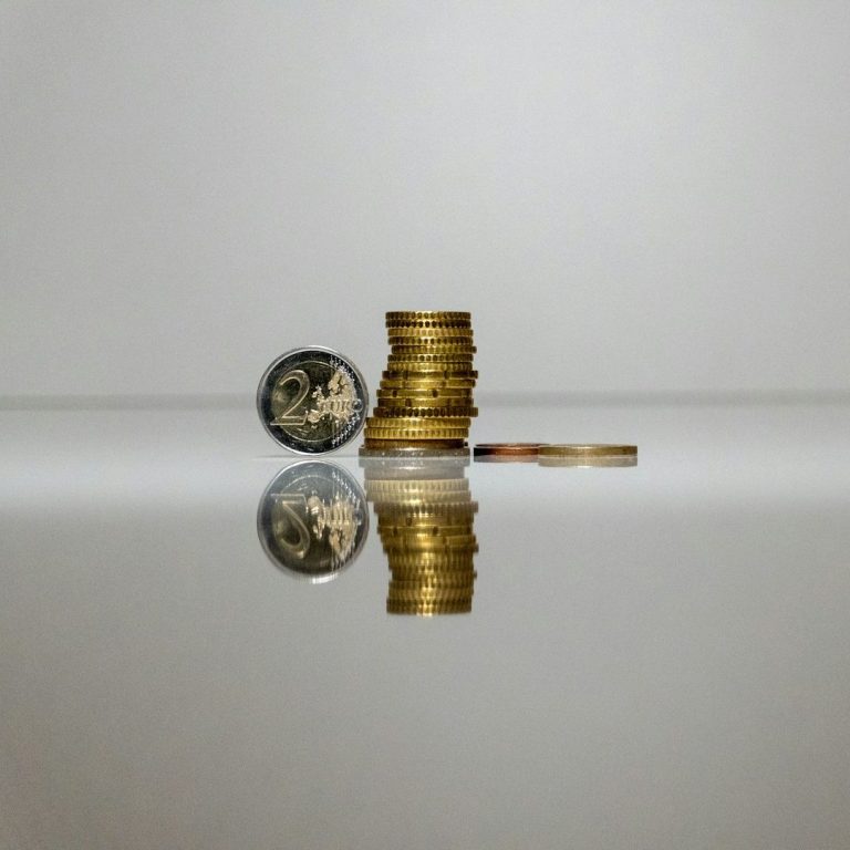A stack of gold coins beside a single silver coin and a banknote, reflecting on a surface.